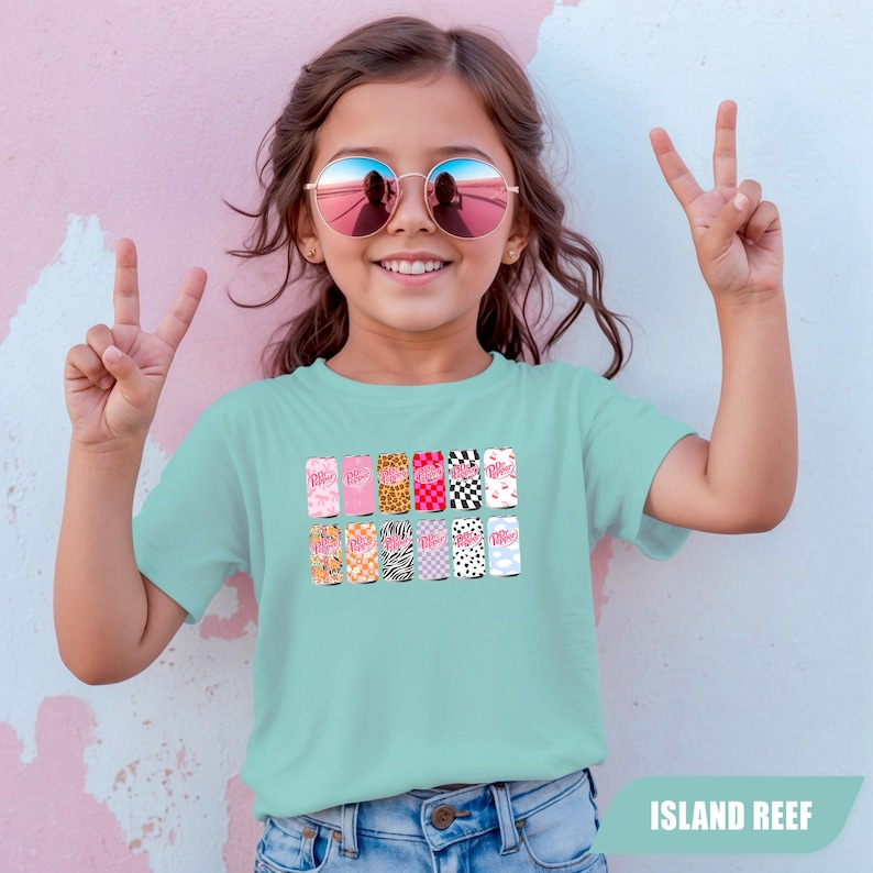 May include: A light mint green short-sleeve t-shirt featuring a graphic print of twelve soda cans arranged in two rows. Each can displays a unique pattern, including leopard print, zebra stripes, checkerboard, and floral designs, with the Dr Pepper logo.