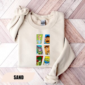 May include: A sand-colored crewneck sweatshirt featuring a graphic print of Toy Story characters including Bo Peep, Mr. Potato Head, Buzz Lightyear, Woody, Rex, and an Alien. The design includes the text 30 YEARS in green and blue lettering.