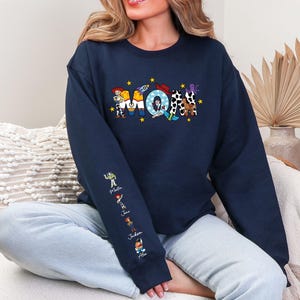 Op de afbeelding: Marineblauwe sweatshirt met het woord "MOM" in kleurrijke, cartoonachtige letters. De letters zijn versierd met personages uit een populaire animatiefilm. De linkermouw heeft extra karakterillustraties en namen.