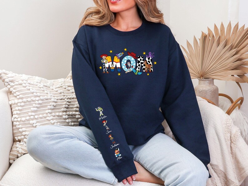 Op de afbeelding: Marineblauwe sweatshirt met het woord "MOM" in kleurrijke, cartoonachtige letters. De letters zijn versierd met personages uit een populaire animatiefilm. De linkermouw heeft extra karakterillustraties en namen.