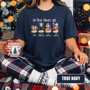 May include: A navy blue t-shirt with the text "IN THIS HOUSE WE" above four cartoon characters. Below the characters are the words "LOVE FAMILY", "LOVE THE PUMPKIN KING", "LOVE HALLOWEEN", and "LOVE CHRISTMAS". The words "TRUE NAVY" are at the bottom.