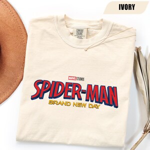 Może przedstawiać: Kremowa koszulka z logo Spider-Man Marvel Studios w kolorze czerwonym i niebieskim oraz złotymi słowami "Brand New Day". Koszulka jest wyprodukowana przez Comfort Colors.