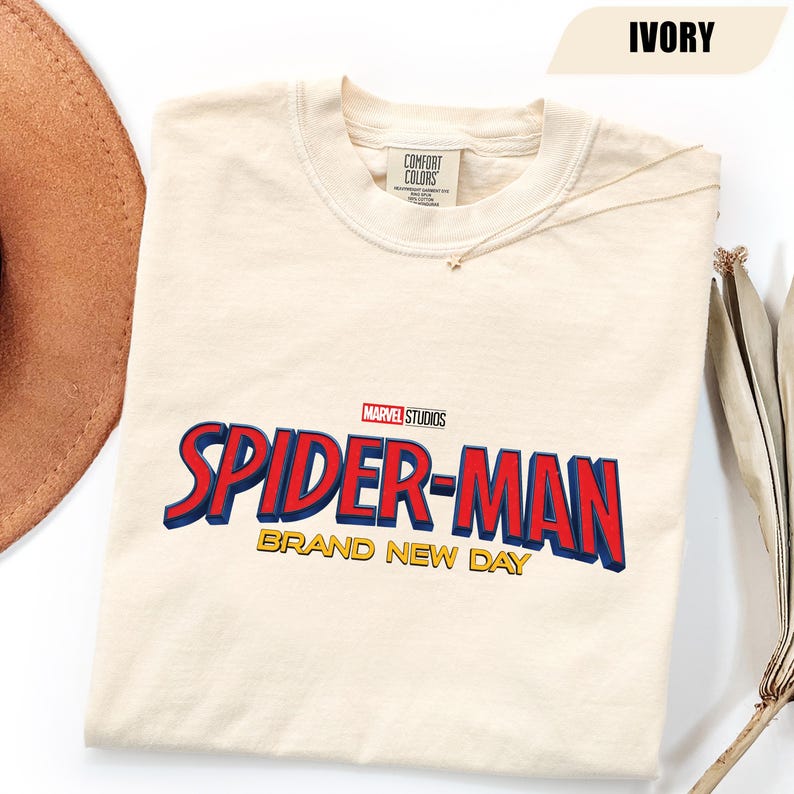 Może przedstawiać: Kremowa koszulka z logo Spider-Man Marvel Studios w kolorze czerwonym i niebieskim oraz złotymi słowami "Brand New Day". Koszulka jest wyprodukowana przez Comfort Colors.