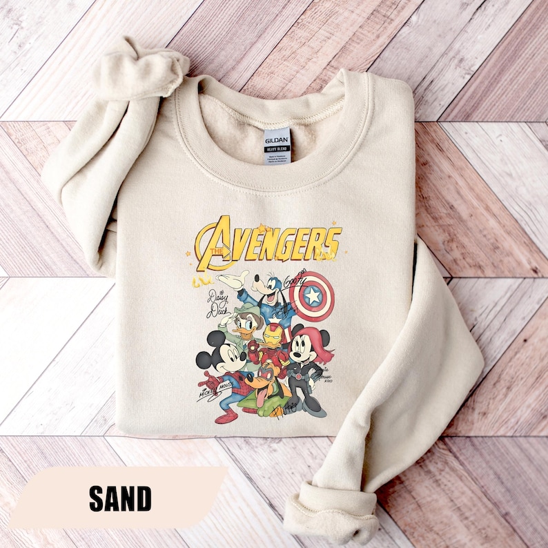 May include: A sand-colored crewneck sweatshirt featuring a graphic print of Disney characters dressed as Avengers. The text reads The Avengers, with illustrations of Mickey Mouse, Donald Duck, Goofy, and Minnie Mouse in superhero costumes and signatures.