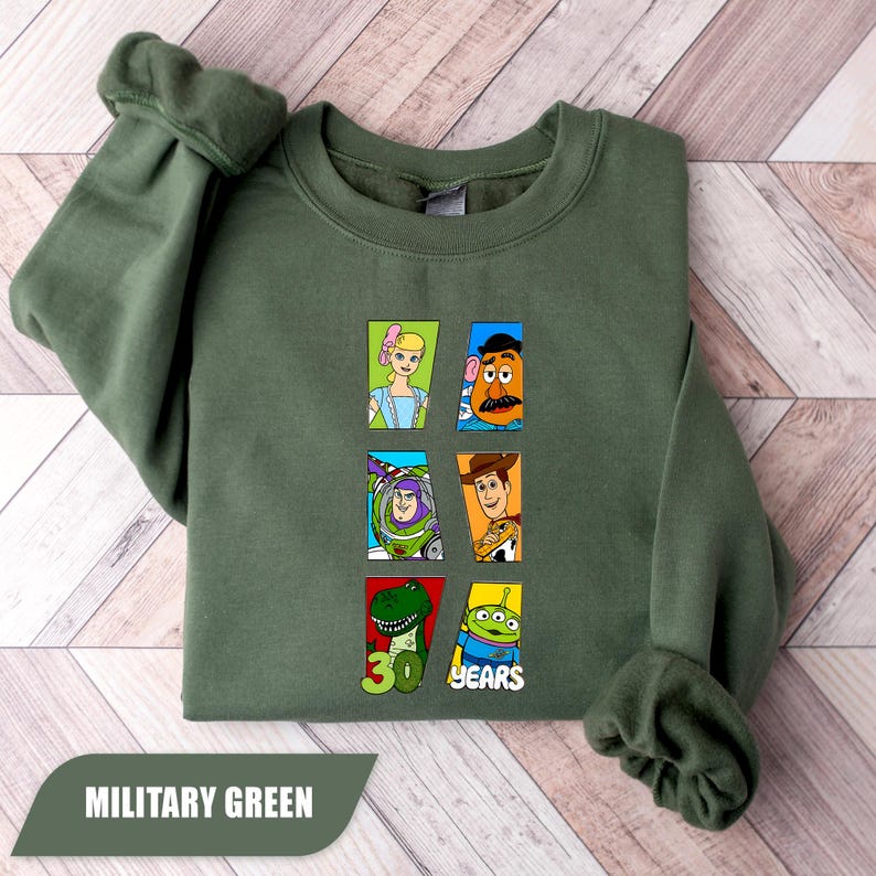 May include: An olive green crewneck sweatshirt featuring a graphic print of six Toy Story characters arranged in rectangular frames. The design includes Bo Peep, Mr. Potato Head, Buzz Lightyear, Woody, Rex, and an Alien, with the text 30 YEARS.