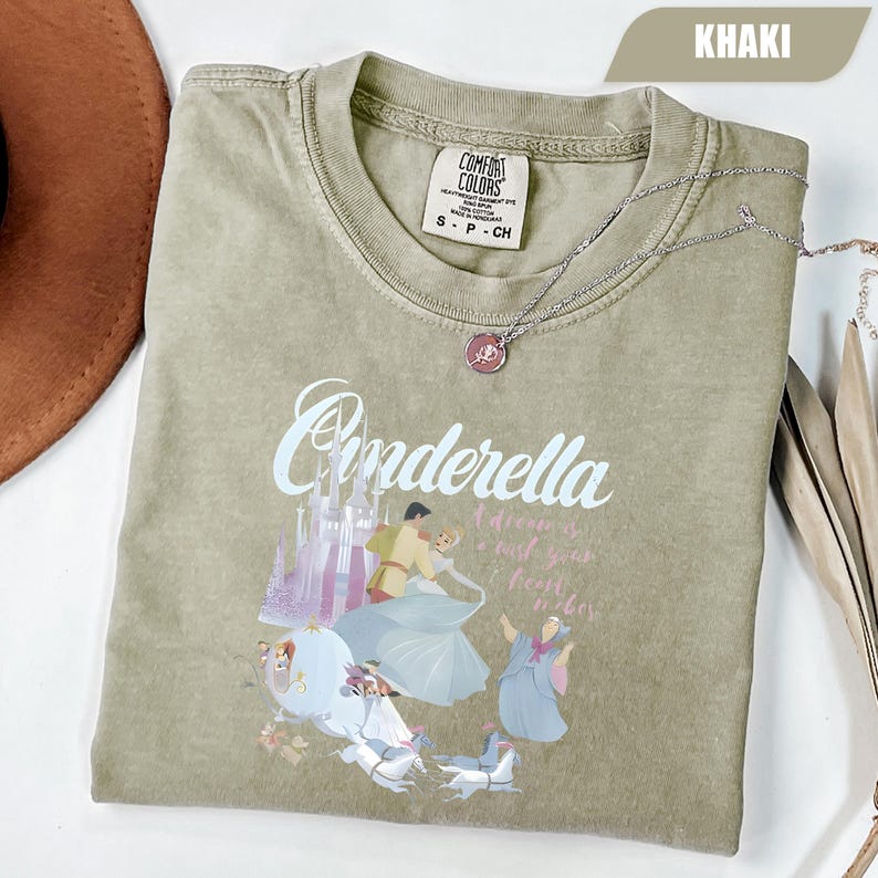 Peut inclure: T-shirt kaki avec le mot "Cinderella" en &eacute;criture cursive, et un dessin de Cendrillon et du Prince Charmant dansant. Le t-shirt a &eacute;galement un dessin d'un ch&acirc;teau et d'un carrosse. Un collier argent&eacute; avec un pendentif rose est sur le t-shirt.