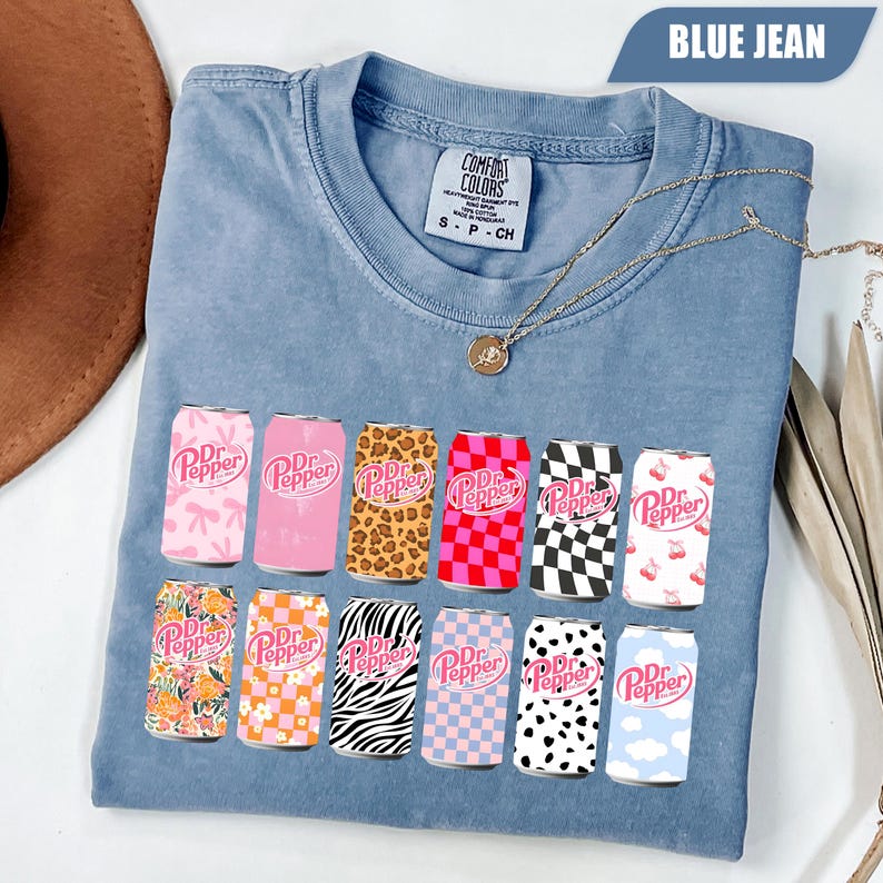 May include: A blue jean colored t-shirt with a Comfort Colors label at the neck. The front features a graphic print of twelve soda cans, each with a unique pattern including leopard print, checkerboard, floral, zebra stripes, and clouds.