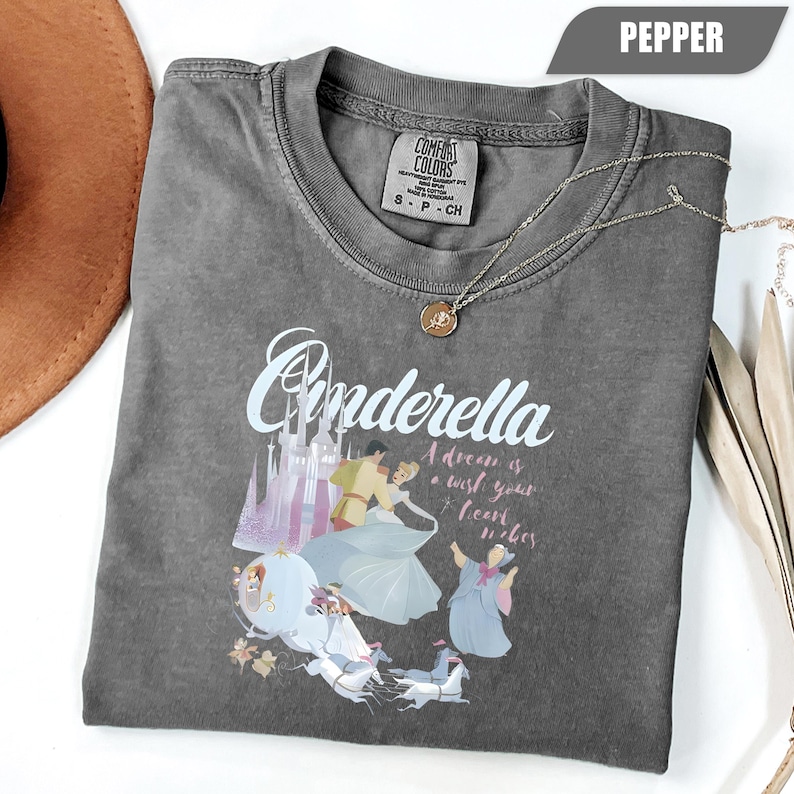 Peut inclure: T-shirt gris avec le mot "Cinderella" et une illustration de conte de f&eacute;es. Le texte "A dream is a wish your heart makes" est &eacute;galement visible. Le t-shirt est de couleur poivre.