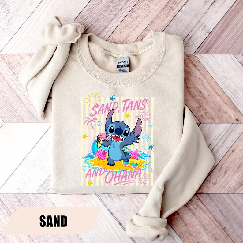May include: A sand-colored crewneck sweatshirt featuring a graphic of the blue character Stitch holding an ice cream cone on a beach. The design includes the text "SAND, TANS AND OHANA" surrounded by sun, palm tree, shell, and star illustrations.