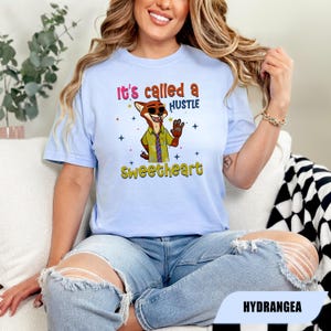 K&ouml;nnte beinhalten: Hellblaues T-Shirt mit einem Cartoon-Fuchs-Charakter und dem Text "It's called a HUSTLE Sweetheart". Der Fuchs tr&auml;gt ein Hemd und eine Krawatte. Das Design enth&auml;lt Sterne und ist auf einem hellblauen T-Shirt gedruckt.