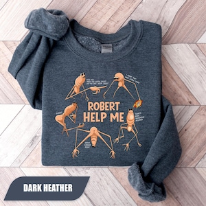 May include: Dark heather sweatshirt with a humorous graphic. The design features several cartoonish creatures with the text "ROBERT HELP ME" and speech bubbles. The sweatshirt is folded, showing the dark blue fabric and the graphic.