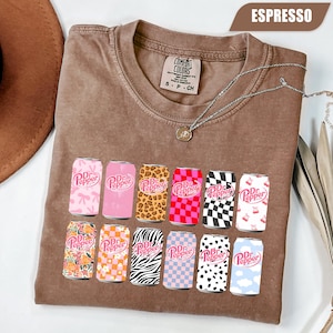 May include: An espresso-colored crew neck t-shirt featuring a graphic print of twelve soda cans, each with a unique pattern including leopard print, checkerboard, floral, zebra stripes, and cow print, all displaying the Dr Pepper logo.