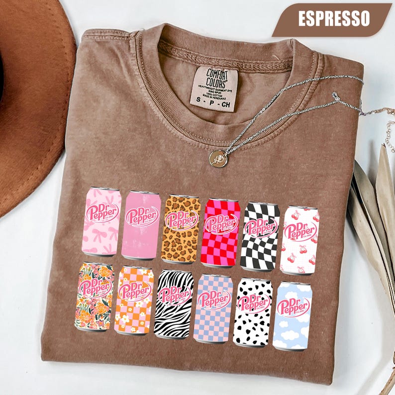 May include: An espresso-colored crew neck t-shirt featuring a graphic print of twelve soda cans, each with a unique pattern including leopard print, checkerboard, floral, zebra stripes, and cow print, all displaying the Dr Pepper logo.