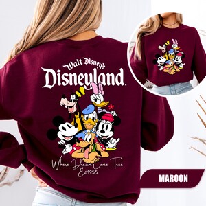 May include: A maroon long-sleeve sweatshirt featuring a large graphic on the back with the text Walt Disney's Disneyland Where Dreams Come True Est 1955 and illustrations of Mickey Mouse, Minnie Mouse, Donald Duck, Daisy Duck, Goofy, and Pluto.