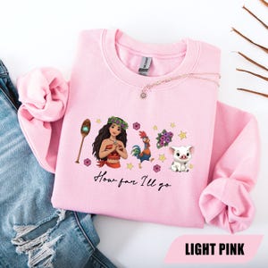 Pu&ograve; includere: Felpa rosa chiaro con una grafica di una donna polinesiana, un gallo, un maiale e la scritta "How far I'll go". Il design include elementi floreali e stelle. La parte inferiore della felpa riporta la scritta "LIGHT PINK".