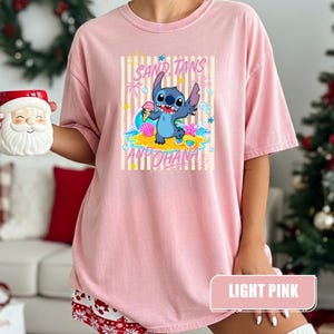 Op de afbeelding: Een lichtroze t-shirt met een afbeelding van het blauwe personage Stitch die een ijshoorntje vasthoudt op een strand. Het ontwerp bevat gele verticale strepen, schelpen, een zeester en de tekst SAND, TANS AND OHANA in roze letters.