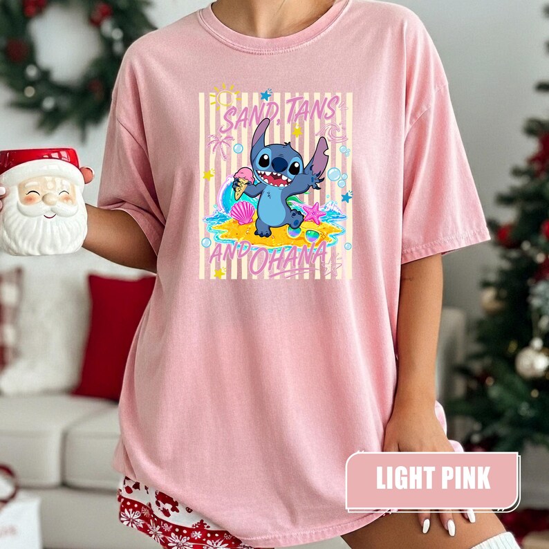 Op de afbeelding: Een lichtroze t-shirt met een afbeelding van het blauwe personage Stitch die een ijshoorntje vasthoudt op een strand. Het ontwerp bevat gele verticale strepen, schelpen, een zeester en de tekst SAND, TANS AND OHANA in roze letters.