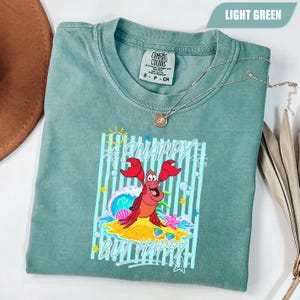 May include: Light green t-shirt with a graphic of a red crab on a sandy beach with the words "Snappy But Happy". The shirt has a crew neck and short sleeves. A silver necklace with a pendant is draped across the shirt.