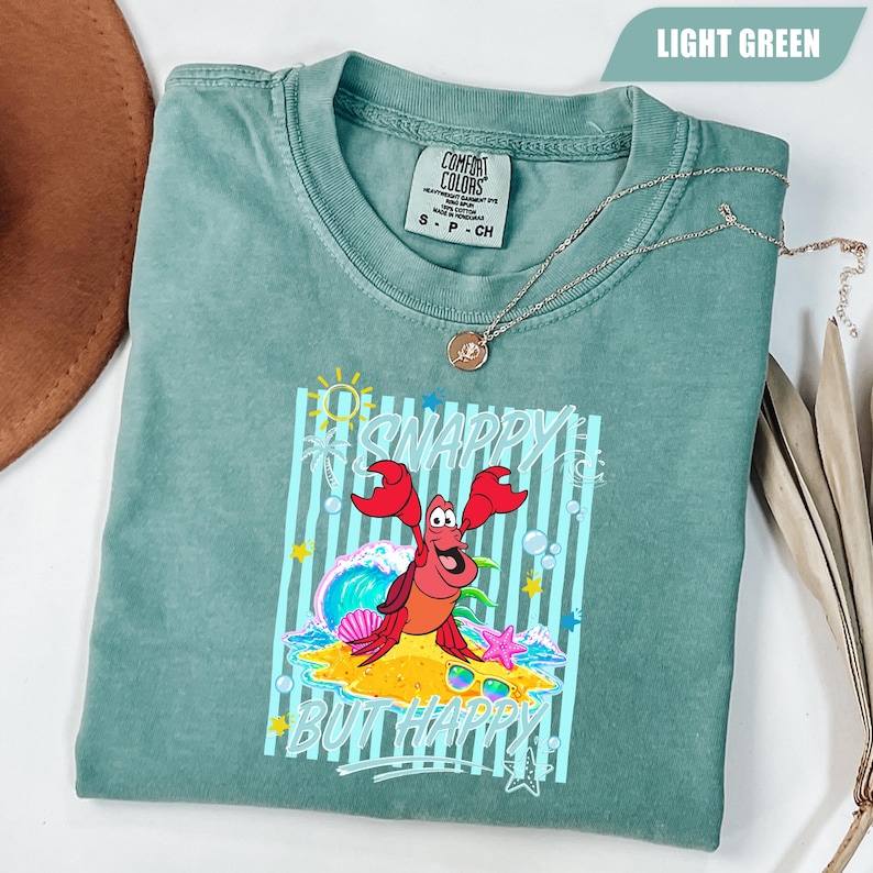 May include: Light green t-shirt with a graphic of a red crab on a sandy beach with the words "Snappy But Happy". The shirt has a crew neck and short sleeves. A silver necklace with a pendant is draped across the shirt.