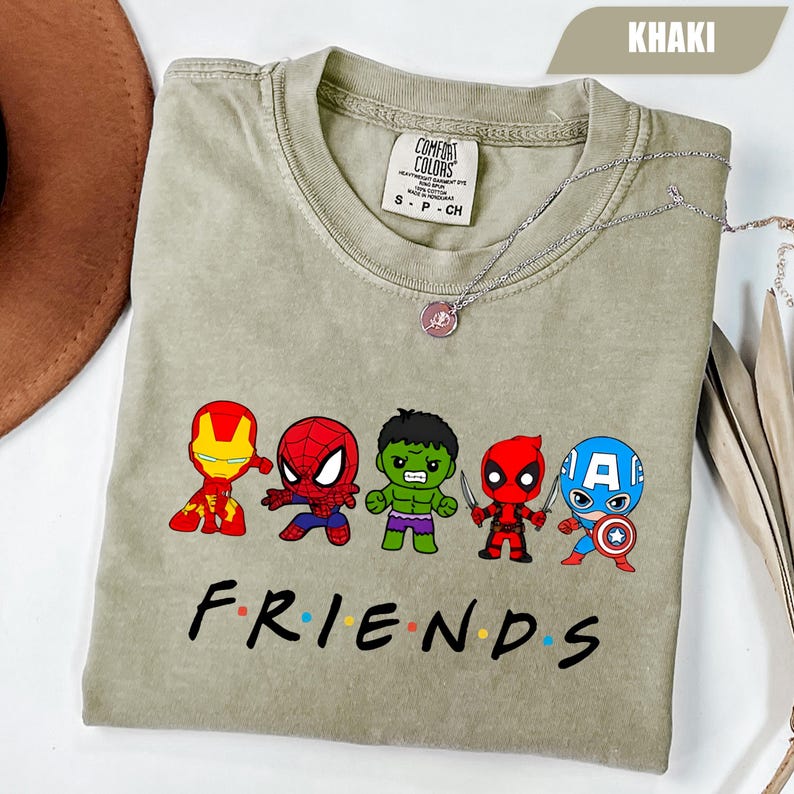 May include: Khaki-colored t-shirt featuring cartoon versions of Iron Man, Spider-Man, Hulk, Deadpool, and Captain America. The word "FRIENDS" is printed below the characters in a colorful font. A silver necklace with a pendant is on the shirt.