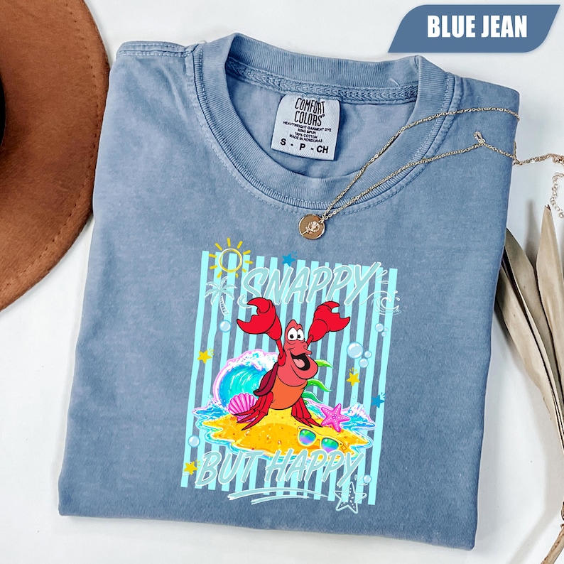 May include: Blue jean Comfort Colors t-shirt featuring a graphic of a red crab with the text "Snappy But Happy". The shirt has a crew neck and short sleeves. A gold necklace is draped across the shirt.