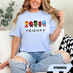 May include: Light blue t-shirt featuring cartoon versions of Iron Man, Spider-Man, Hulk, Deadpool, and Captain America. The word "FRIENDS" is printed below the characters in black. The shirt is made of a soft material.