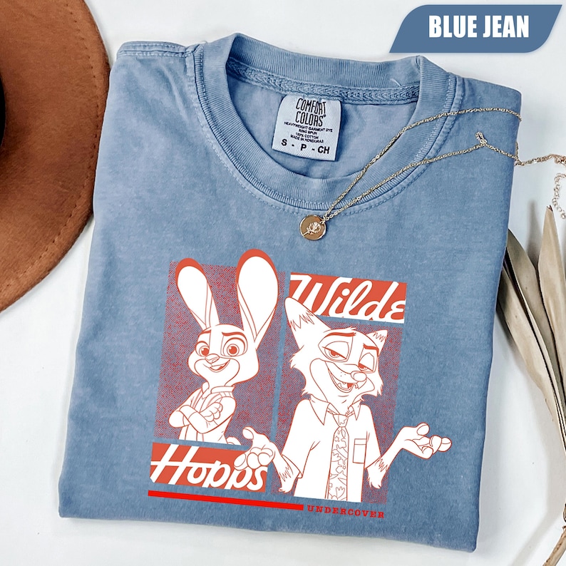 Puede incluir: Camiseta azul vaquero con un gr&aacute;fico rojo y blanco de los personajes de dibujos animados Judy Hopps y Nick Wilde. La camiseta tiene las palabras "Wilde" y "Hopps" en rojo. Un collar dorado est&aacute; drapeado sobre la camiseta.
