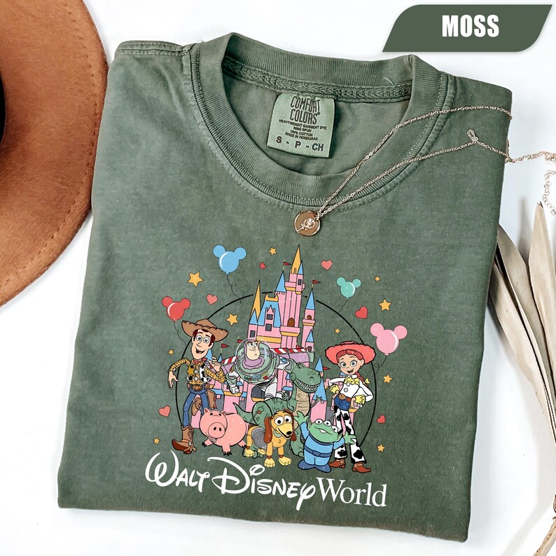 Puede incluir: Camiseta verde musgo con un gr&aacute;fico colorido de personajes de Toy Story y un castillo, con el texto "Walt Disney World". La camiseta tiene cuello redondo y mangas cortas. Un collar plateado con un colgante est&aacute; sobre la camiseta.
