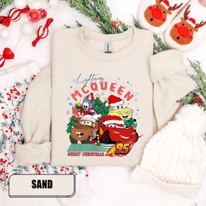 May include: Sand-colored sweatshirt featuring a graphic of Lightning McQueen and other Cars characters wearing Santa hats, with the text "Lightning McQueen" and "Merry Christmas". The number "95" is also visible.