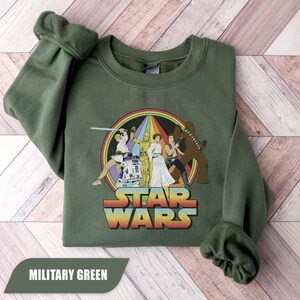 May include: Military green sweatshirt featuring a Star Wars graphic. The design includes characters like Luke Skywalker, Princess Leia, and Chewbacca, set against a rainbow backdrop with the Star Wars logo.