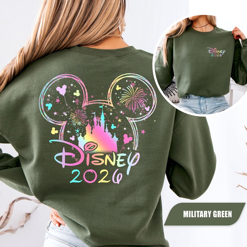 Puede incluir: Sudadera verde militar con un colorido dise&ntilde;o de castillo de Disney y fuegos artificiales. La parte trasera muestra un gran contorno de la cabeza de Mickey Mouse con el texto "Disney 2026". Una versi&oacute;n m&aacute;s peque&ntilde;a del dise&ntilde;o est&aacute; en la parte delantera.