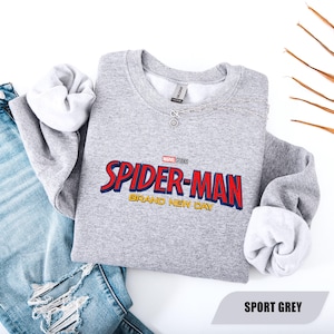 Puede incluir: Una sudadera gris claro con las palabras "SPIDER-MAN" en rojo y azul, y "BRAND NEW DAY" en dorado. La sudadera se combina con unos vaqueros azules y un collar plateado. El texto "SPORT GREY" est&aacute; en la parte inferior.