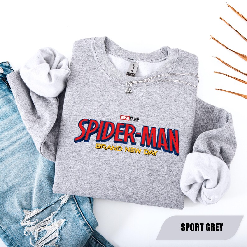 Puede incluir: Una sudadera gris claro con las palabras "SPIDER-MAN" en rojo y azul, y "BRAND NEW DAY" en dorado. La sudadera se combina con unos vaqueros azules y un collar plateado. El texto "SPORT GREY" est&aacute; en la parte inferior.