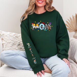 Op de afbeelding: Donkergroene sweatshirt met een kleurrijk grafisch ontwerp. Het ontwerp toont het woord "Mom" met cartoonfiguren en sterren. De linkermouw heeft extra cartoonfiguren en namen. De sweatshirt is een crew neck stijl.