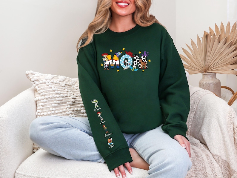 Op de afbeelding: Donkergroene sweatshirt met een kleurrijk grafisch ontwerp. Het ontwerp toont het woord "Mom" met cartoonfiguren en sterren. De linkermouw heeft extra cartoonfiguren en namen. De sweatshirt is een crew neck stijl.
