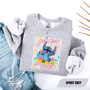 May include: A heather grey crewneck sweatshirt featuring a graphic of the blue character Stitch holding an ice cream cone on a beach. The design includes the text SAND, TANS AND OHANA, along with palm trees, seashells, and stars.