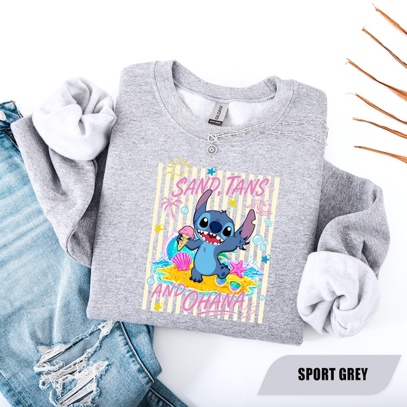 May include: A heather grey crewneck sweatshirt featuring a graphic of the blue character Stitch holding an ice cream cone on a beach. The design includes the text SAND, TANS AND OHANA, along with palm trees, seashells, and stars.
