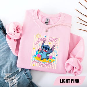 May include: A light pink crewneck sweatshirt featuring a graphic of the blue character Stitch holding an ice cream cone on a beach. The text SAND, TANS AND OHANA is printed in pink above and below the character, surrounded by sun, wave, and shell icons.