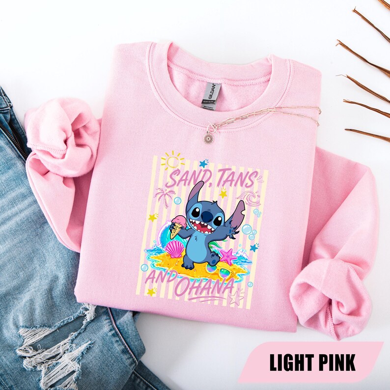 May include: A light pink crewneck sweatshirt featuring a graphic of the blue character Stitch holding an ice cream cone on a beach. The text SAND, TANS AND OHANA is printed in pink above and below the character, surrounded by sun, wave, and shell icons.