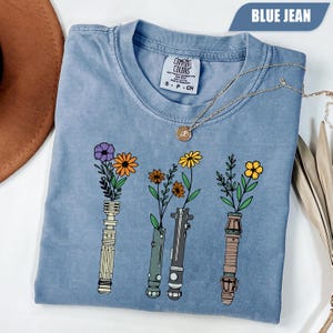 Könnte beinhalten: Ein blaues Jeans-T-Shirt mit floralem Design. Die Grafik zeigt drei Lichtschwertgriffe mit bunten Blumen. Das Shirt hat einen Rundhalsausschnitt und kurze Ärmel. Eine goldene Halskette ist über dem Shirt drapiert.