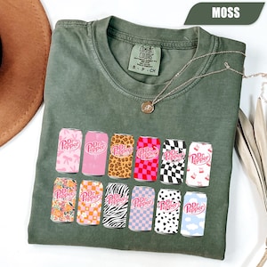 May include: A moss green short-sleeve t-shirt featuring a graphic print of twelve soda cans, each with a unique pattern including leopard print, checkerboard, zebra stripes, floral, and bows. The shirt has a crew neckline and a Comfort Colors label.