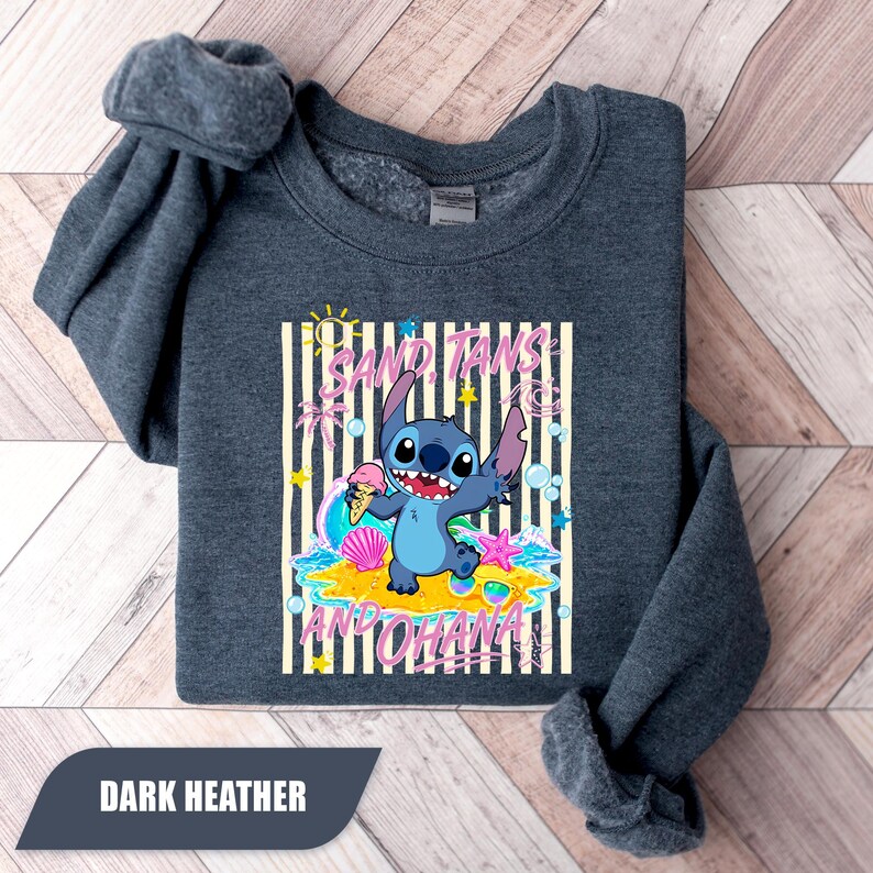 May include: A dark heather gray crewneck sweatshirt featuring a graphic of the blue character Stitch holding an ice cream cone on a beach. The design includes the text SAND, TANS AND OHANA, surrounded by palm trees, a seashell, and a starfish.
