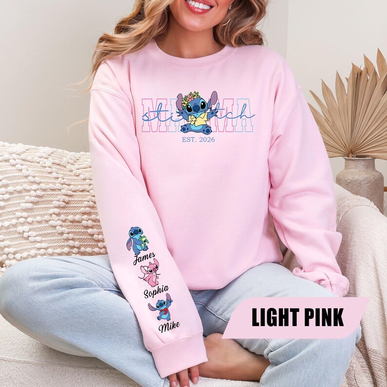 May include: A light pink long-sleeve sweatshirt featuring a graphic of the character Stitch wearing a flower crown. The text reads MAMA and EST. 2026. The sleeve displays three small character illustrations with the names James, Sophia, and Mike.
