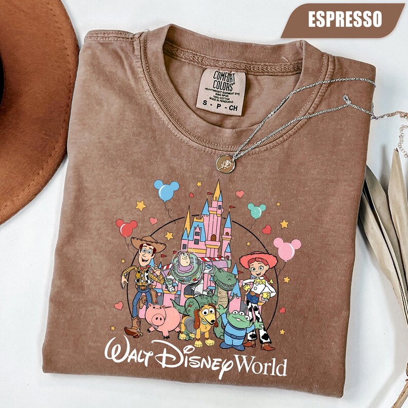 Puede incluir: Camiseta color espresso con un gr&aacute;fico de personajes de Disney frente a un castillo. La camiseta tiene cuello redondo y mangas cortas. Un collar plateado con un colgante est&aacute; drapeado sobre la camiseta. El texto "Walt Disney World" est&aacute; impreso en la parte inferior.