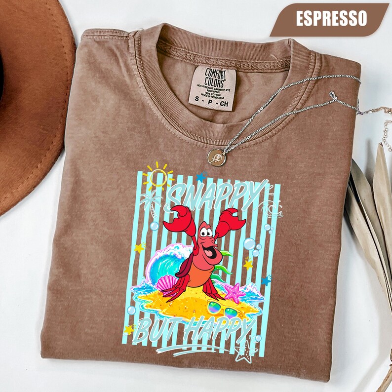 May include: Espresso-colored t-shirt with a graphic of a cartoon crab on a sandy beach with the words "Snappy But Happy". The shirt has a crew neck and short sleeves. A silver necklace is draped across the shirt.