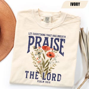 Puede incluir: Camiseta de color marfil con las palabras "PRAISE THE LORD" en azul marino, acompañada de un ramo de flores. El texto "LET EVERYTHING THAT HAS BREATH" está encima de "PRAISE", y "PSALM 150:6" debajo. La camiseta tiene cuello redondo.