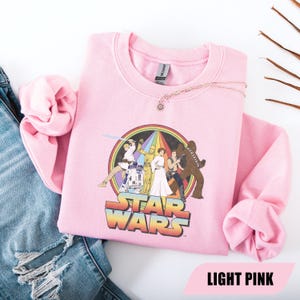 May include: Light pink Star Wars sweatshirt featuring a colorful graphic of characters from the film series. The graphic includes a rainbow and the words "STAR WARS" in a retro font. The sweatshirt is made of a soft material.