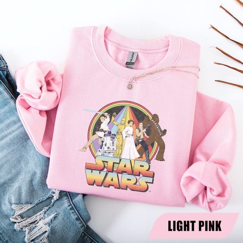 May include: Light pink Star Wars sweatshirt featuring a colorful graphic of characters from the film series. The graphic includes a rainbow and the words "STAR WARS" in a retro font. The sweatshirt is made of a soft material.