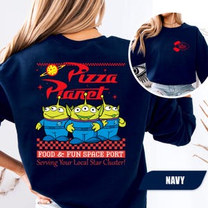 Puede incluir: Sudadera azul marino con el logo de Pizza Planet y tres personajes alien&iacute;genas verdes. El dise&ntilde;o trasero incluye el texto "FOOD & FUN SPACE PORT Serving Your Local Star Cluster!". Un logotipo m&aacute;s peque&ntilde;o de Pizza Planet est&aacute; en la parte delantera.
