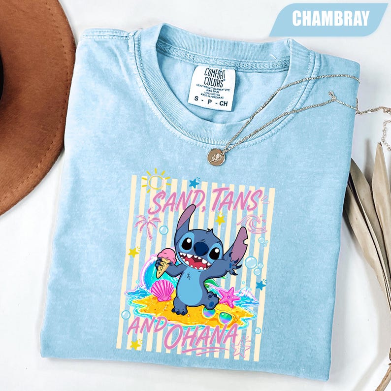 Op de afbeelding: Een lichtblauw t-shirt met korte mouwen en een afbeelding van het personage Stitch die een ijshoorntje vasthoudt op een strand. De tekst luidt SAND, TANS AND OHANA. Het shirt heeft een Comfort Colors-label in de nek en de kleur chambray.